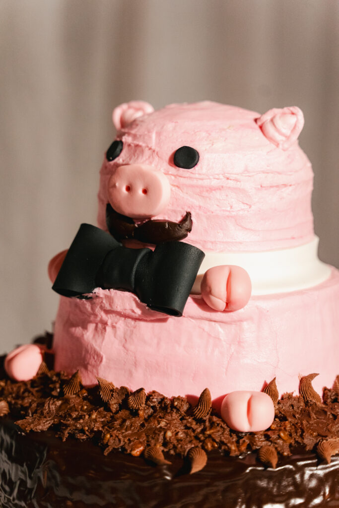 Whimsical pink groom’s cake shaped like a pig wearing a black bow tie with chocolate “mud” icing at the base.
