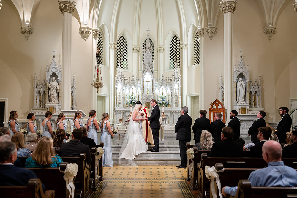 Wedding ceremony inside a cathedral with couple exchanging vows at the altar surrounded by guests.