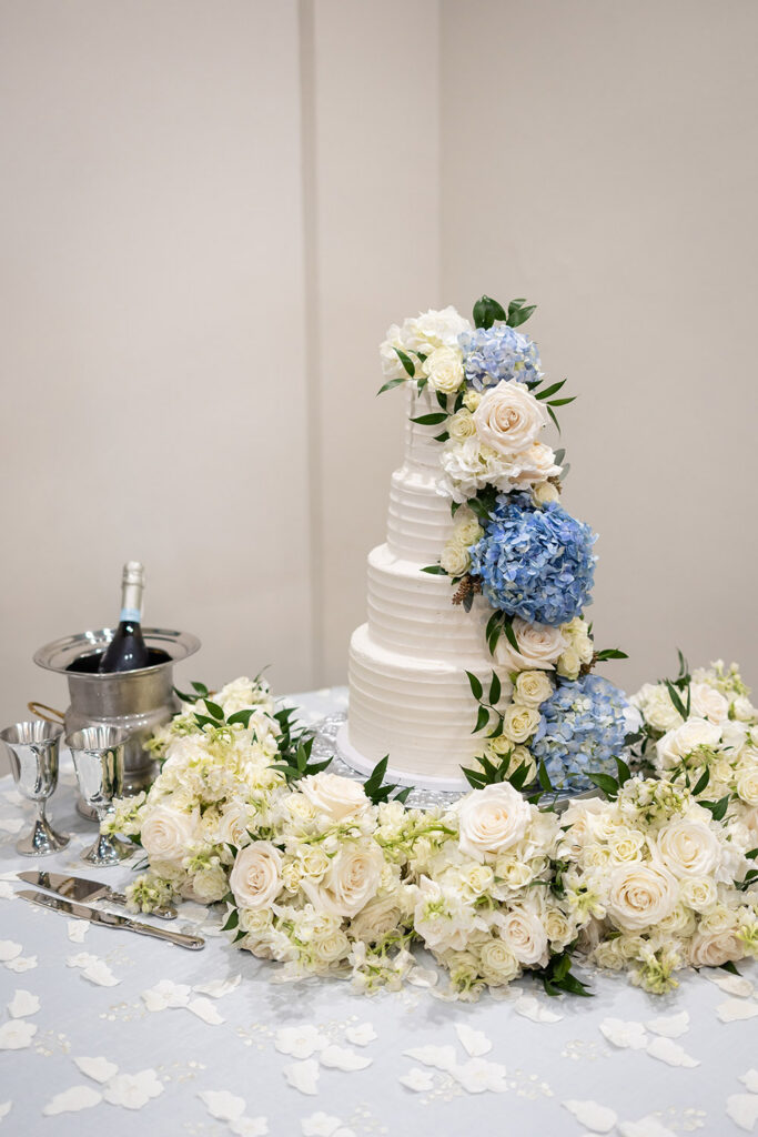 Three-tier white wedding cake decorated with blue hydrangeas and white roses on a reception table.