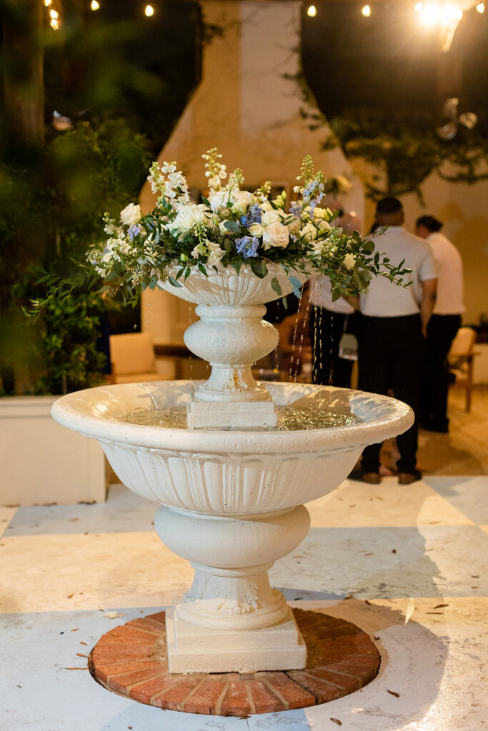 Outdoor fountain decorated with white and blue floral arrangement during evening wedding reception.