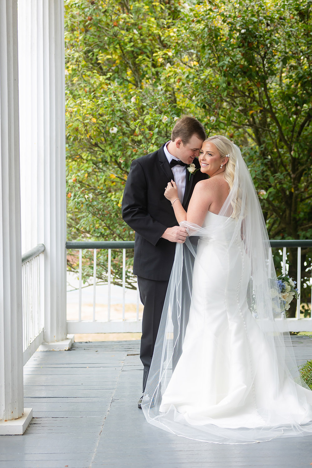 Couple embracing on a porch with greenery backdrop during wedding portraits.