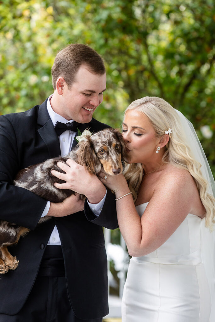 Couple holding a dachshund and kissing the dog during wedding portraits.