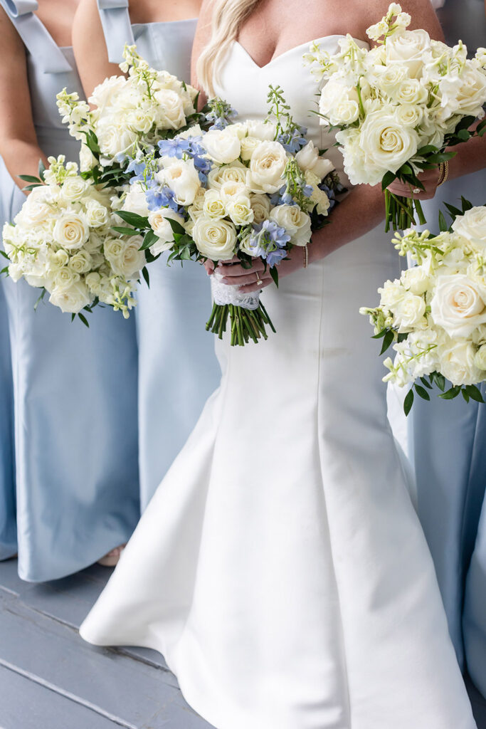 Wedding party holding white and soft blue bouquets with roses and delphinium against pastel blue dresses.