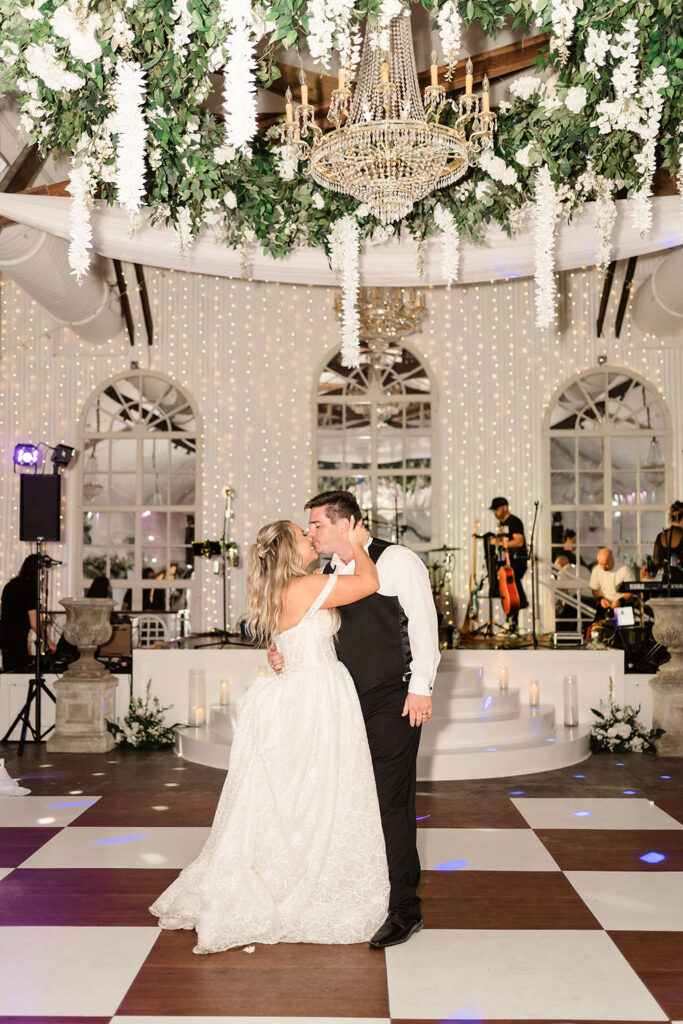 Couple sharing a kiss during their first dance beneath a chandelier and cascading floral reception décor.