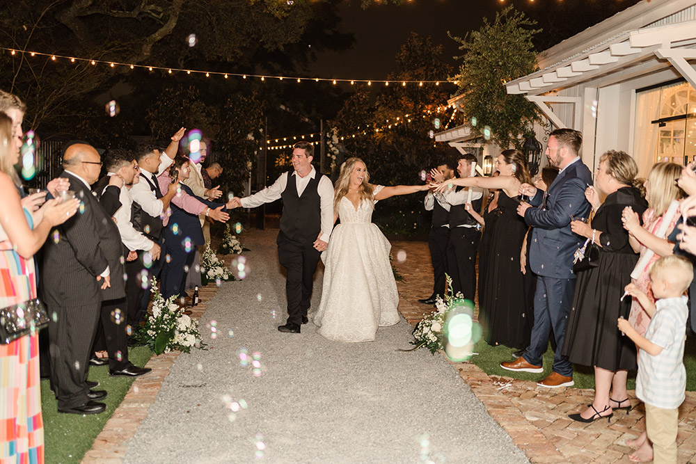 Newlyweds walking through guests blowing bubbles during a nighttime wedding send-off outside the venue.