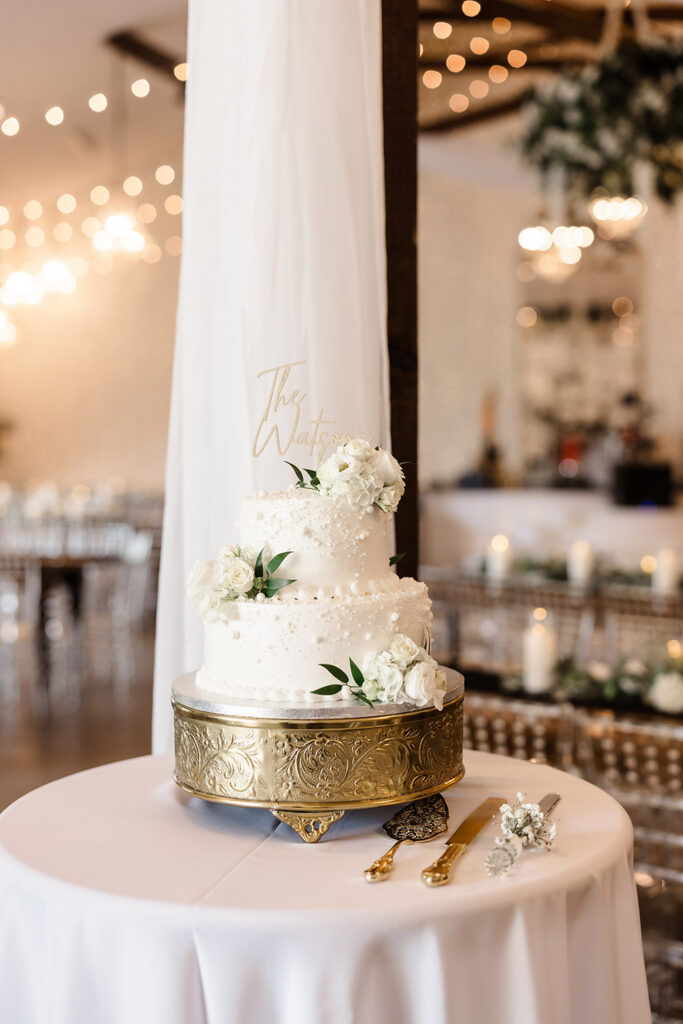 Two-tier white wedding cake decorated with flowers and greenery on a gold stand inside reception hall.