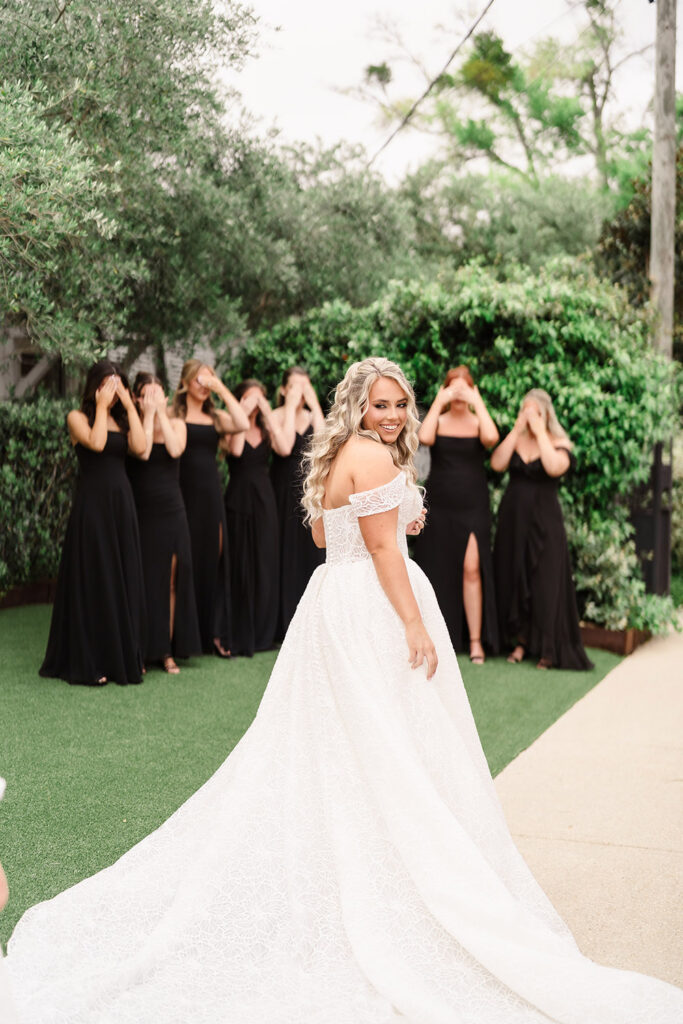 Bride posing in a lace wedding gown while bridesmaids in black dresses stand behind with their eyes covered.