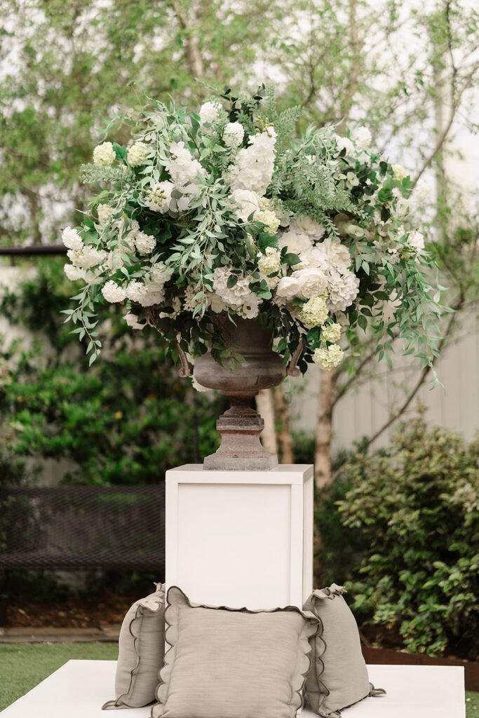 Large white and greenery floral arrangement in a stone urn displayed on a pedestal at outdoor wedding ceremony.