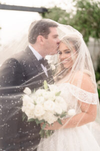 Wedding portrait of couple beneath a veil as one partner kisses the other’s forehead while holding a white bouquet.
