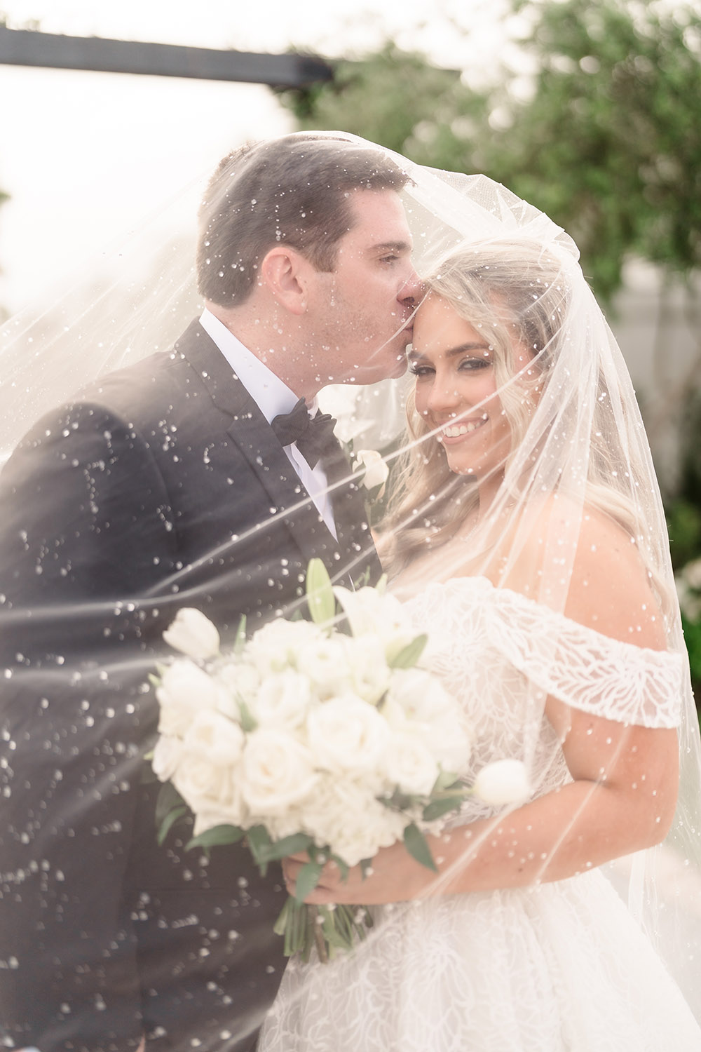 Wedding portrait of couple beneath a veil as one partner kisses the other’s forehead while holding a white bouquet.