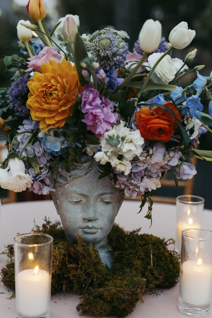 Sculptural centerpiece with colorful flowers arranged in a decorative bust vase surrounded by candles.