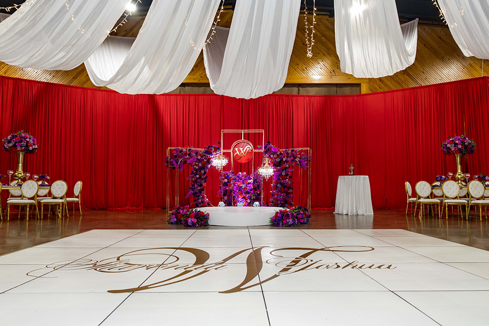 Reception ballroom reveal with red draped walls, suspended white fabric ceiling, floral ceremony stage, and custom monogram dance floor.
