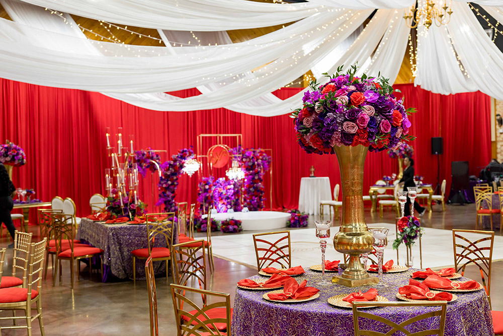 Reception ballroom featuring red drapery, white ceiling swags, gold chairs, and dramatic purple and red floral centerpieces.