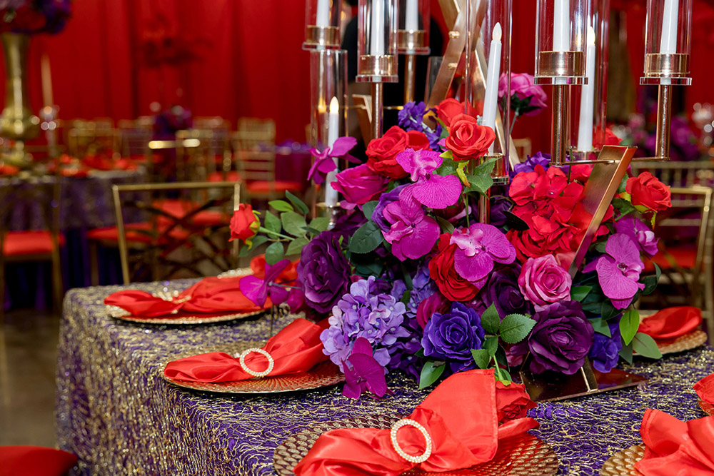 Vibrant reception table featuring purple and red florals, gold candle holders, and red satin napkins on textured linens.