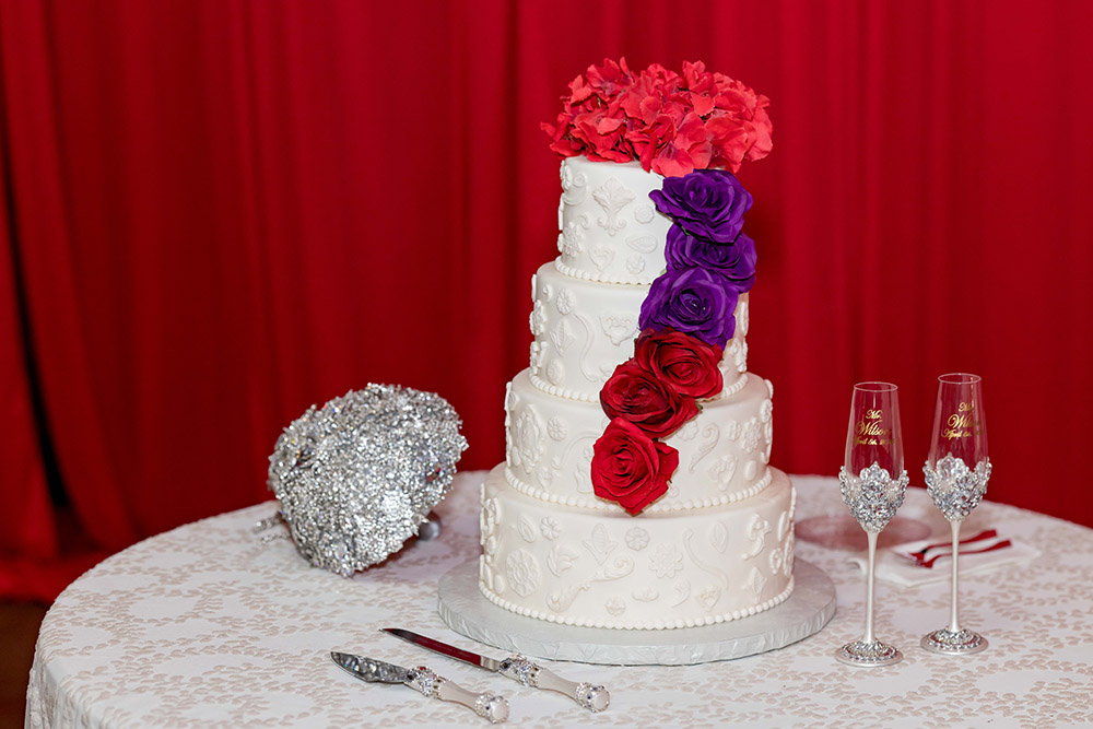 Four-tier white wedding cake adorned with red and purple roses displayed beside crystal champagne flutes.