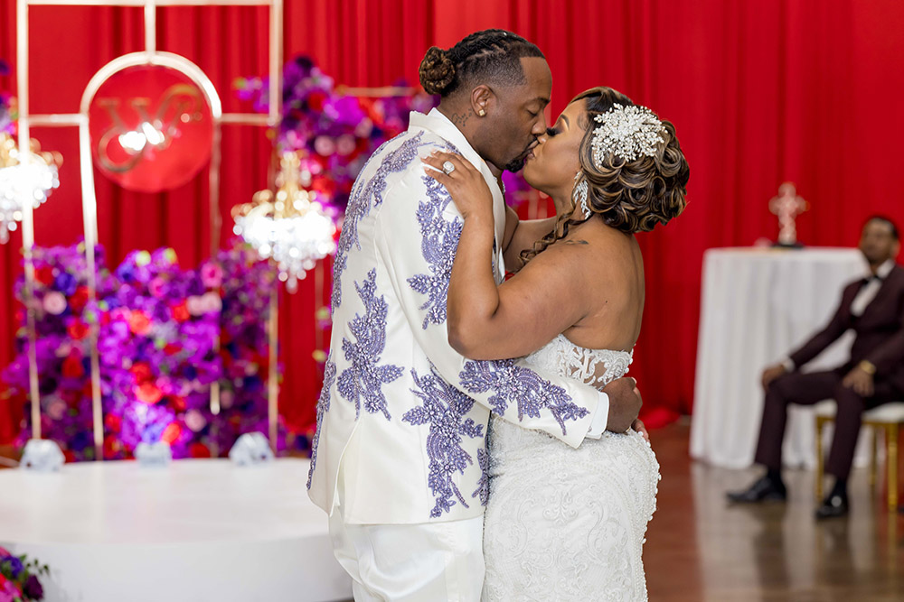 Bride and groom sharing a kiss on the dance floor beneath red drapery and jewel-toned floral décor.