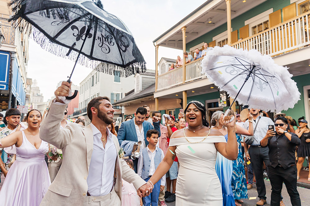 New Orleans second line wedding parade with bride and groom dancing through French Quarter holding umbrellas surrounded by guests
