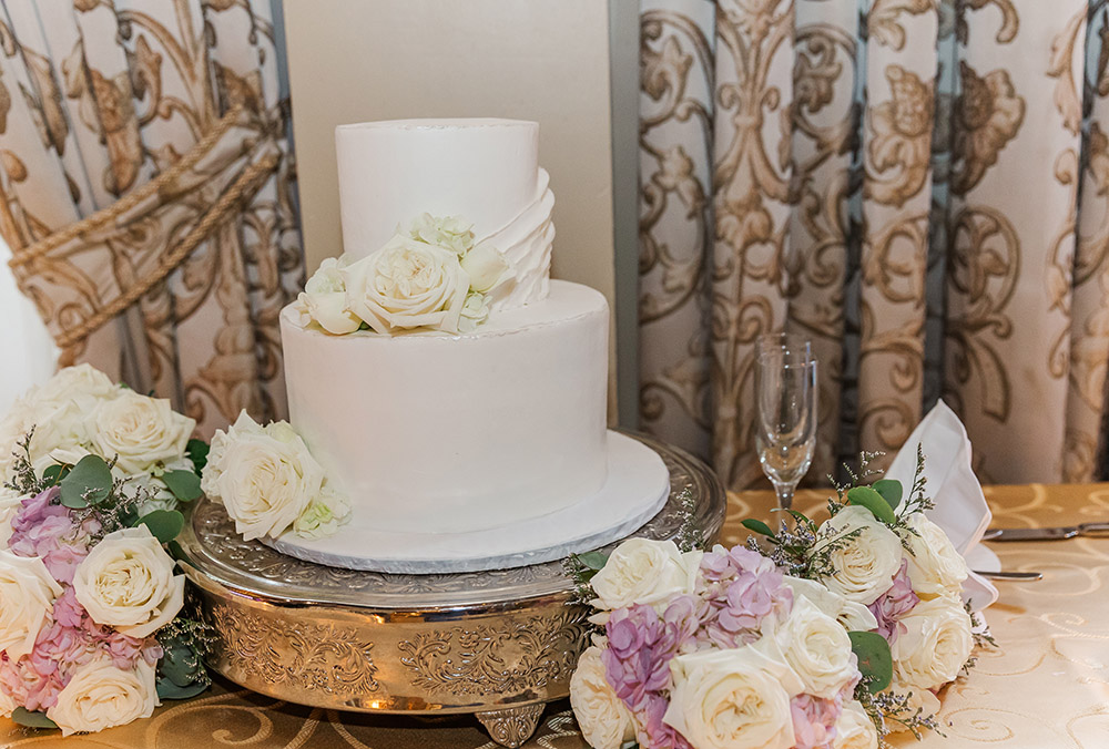 Two-tier white wedding cake with floral decorations on ornate stand surrounded by blush and ivory flowers