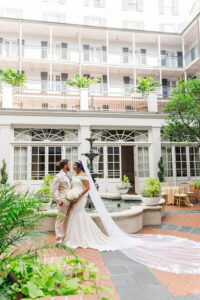 Bride and groom portrait in New Orleans courtyard with fountain and greenery, bride wearing long cathedral veil and holding bouquet
