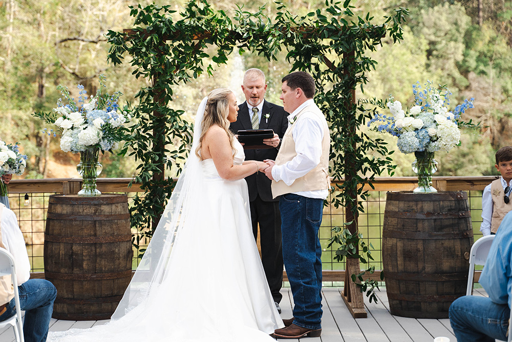 Outdoor wedding ceremony with bride and groom exchanging vows beneath a greenery arbor, surrounded by florals and rustic barrels