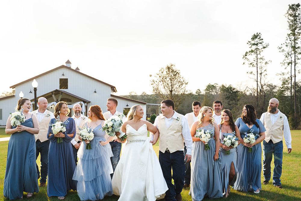 Bride and groom walk with bridesmaids and groomsmen on a sunny lawn in front of a white barn, smiling together in a wedding party. (informative)