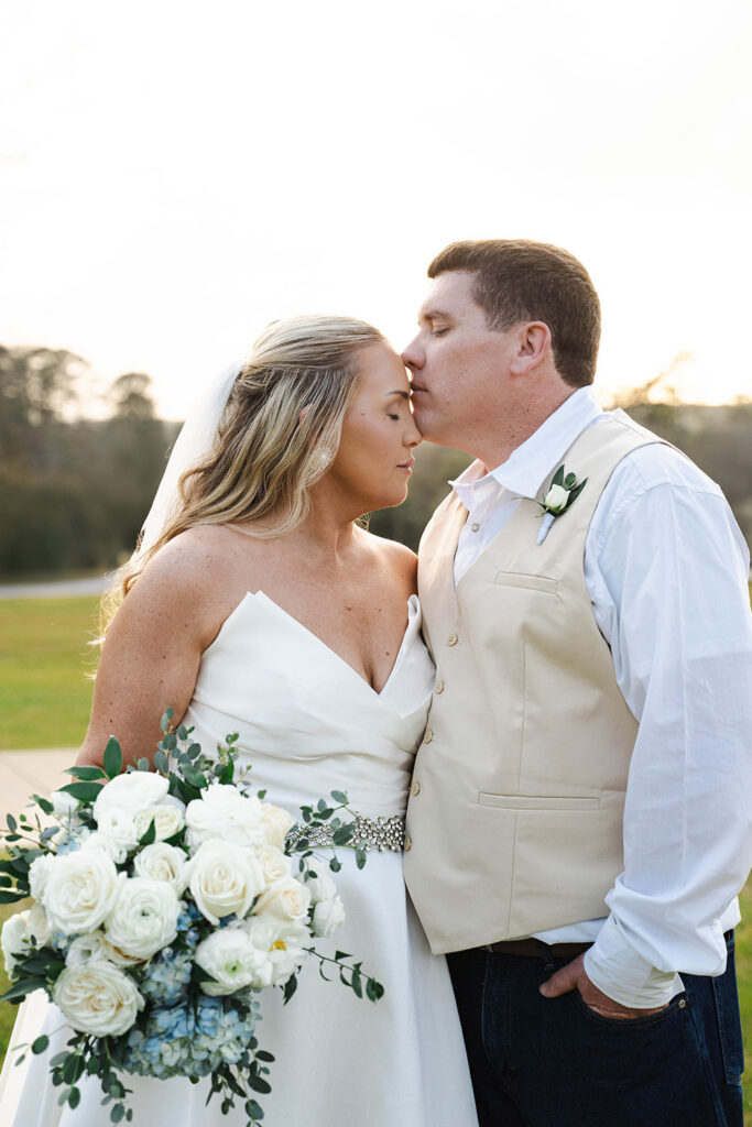 Bride and groom share a kiss with eyes closed outdoors at sunset; she holds a white bouquet of roses and greenery while wearing a white dress and belt.