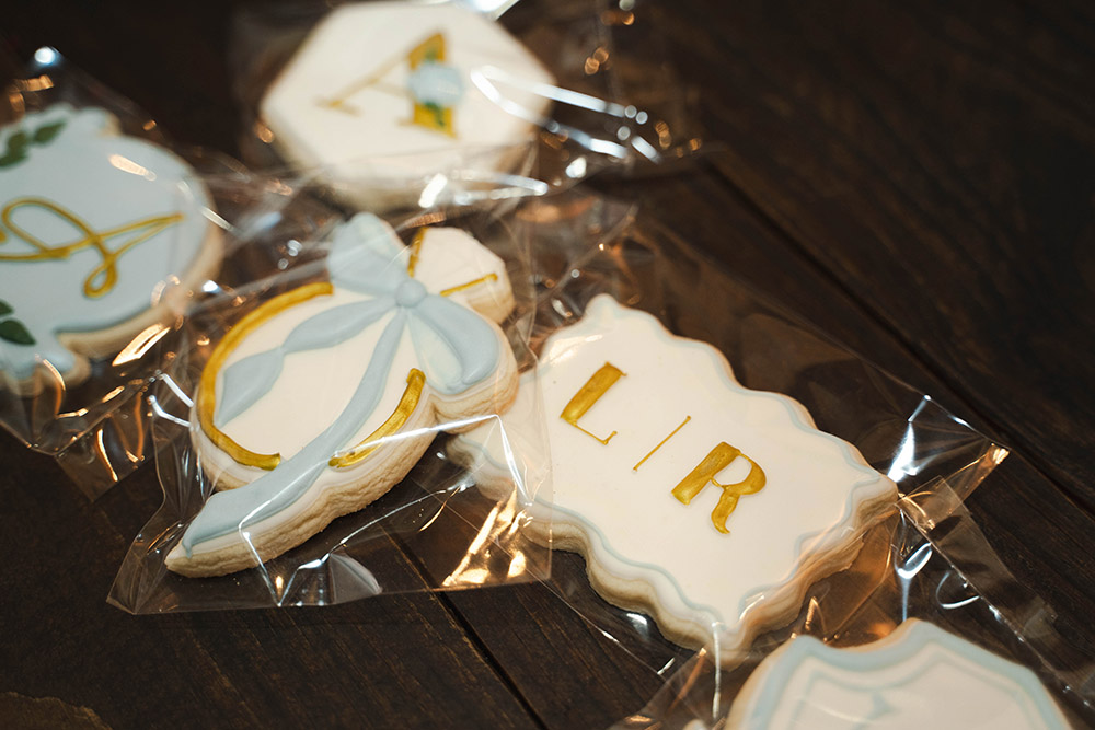 Assorted iced cookies in clear bags on a dark wooden table; one rectangular cookie has gold letters reading 'LIR' and others feature pastel blue bows and designs on white icing.