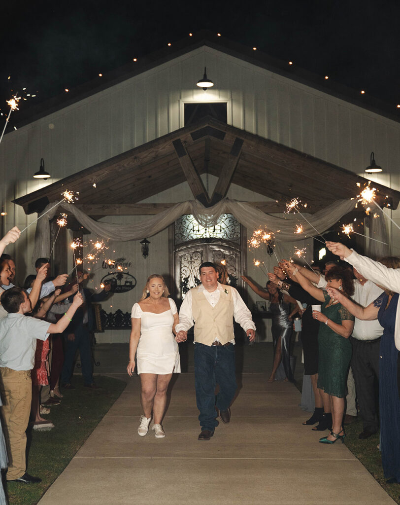 Bride and groom walk hand in hand down a lit outdoor aisle as guests spark firework sparklers on both sides.