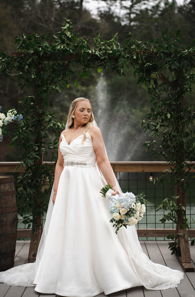 Bride in a white wedding gown with a jeweled belt, holding a pale bouquet, standing beneath a lush green leafy arch on a wooden deck with a fountain in the background.