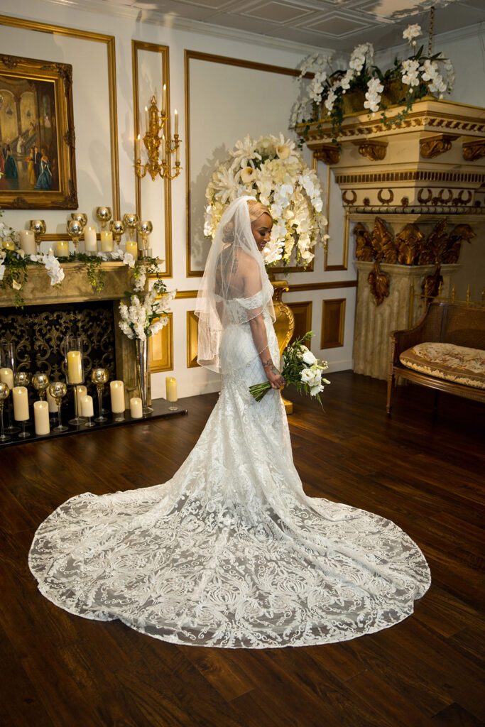 Bride in fitted lace wedding gown with long train and veil holding bouquet in elegant room with gold trim, candles, and white florals