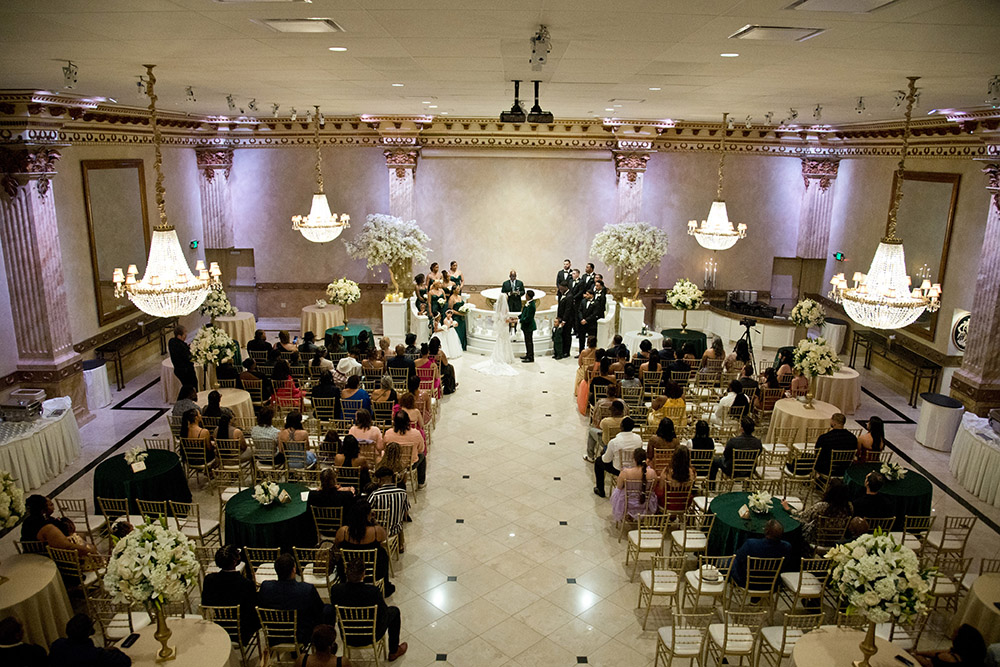 Wide overhead view of grand ballroom wedding ceremony with chandeliers, round guest tables, and floral arrangements framing the aisle