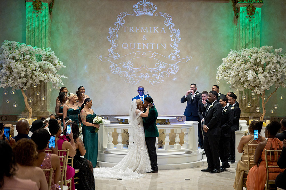 Couple shares first kiss during elegant ballroom wedding ceremony with green uplighting and floral arrangements, monogram reading Tremika and Quentin