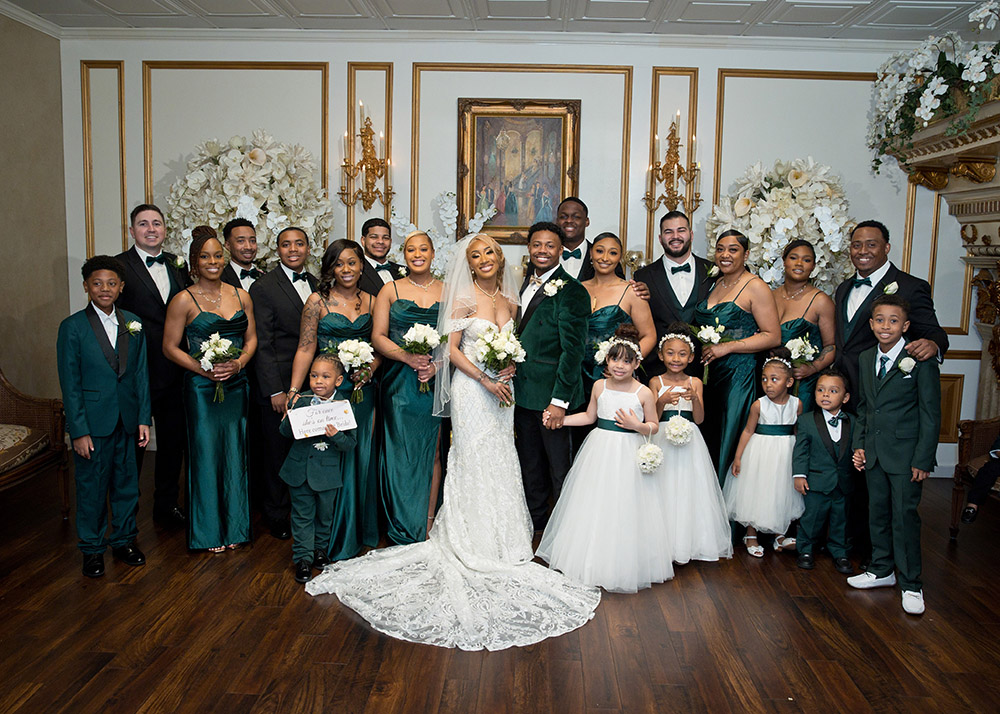 Full wedding party portrait with bride, groom, bridesmaids in emerald dresses, groomsmen in black tuxedos, and flower girls in white dresses