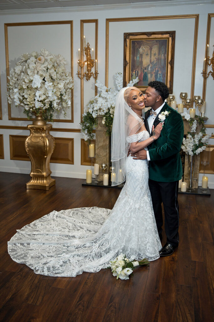 Bride and groom portrait in ornate reception room with gold accents, white florals, and candlelit fireplace as groom kisses bride’s cheek