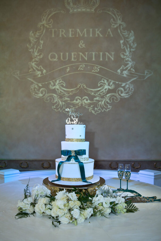 Four-tier white wedding cake with emerald ribbon and gold detailing displayed beneath custom Tremika and Quentin monogram projection