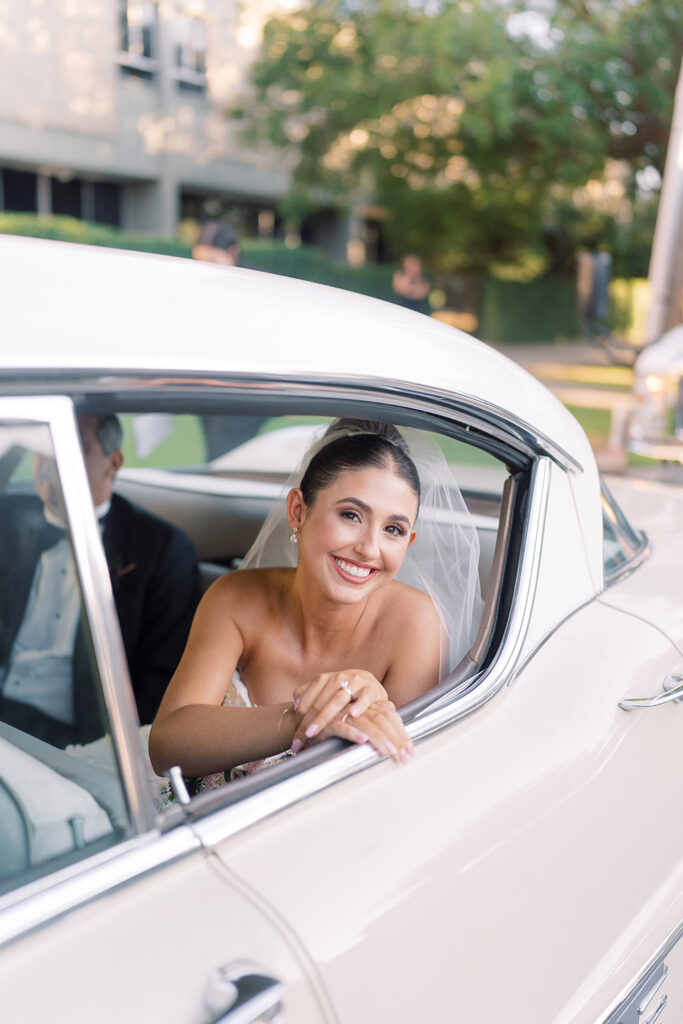 Smiling bride leans out of the backseat window of a vintage white getaway car before the ceremony.