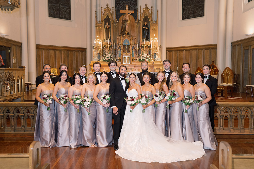 Full wedding party poses inside a historic church with vaulted ceilings, floral arrangements, and polished wood floors.