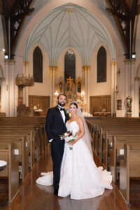 Newlywed couple poses in the center aisle of a grand church, the bride holding a bouquet while her lace train trails behind her.