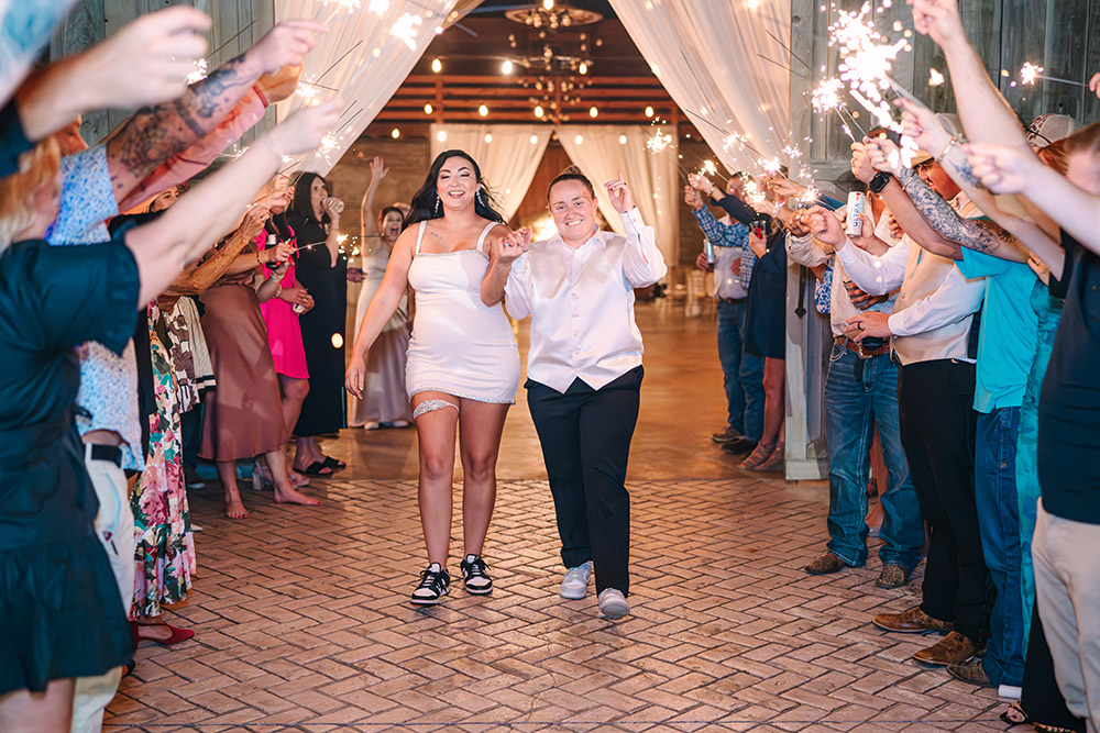 Newlywed brides making a joyful sparkler exit through cheering wedding guests inside a rustic barn venue at the end of the reception.