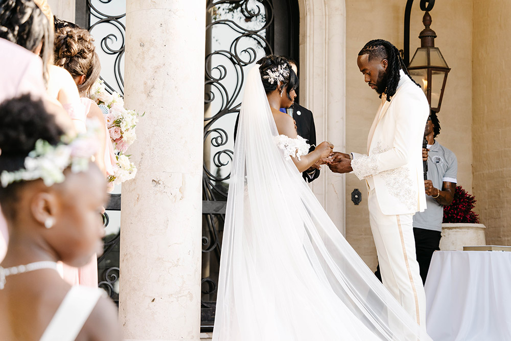 Bride and groom exchange rings during an outdoor wedding ceremony beside marble columns and flowing cathedral-length veil.