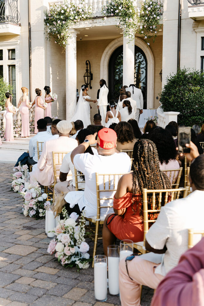 Guests watch as the bride and groom exchange vows during an outdoor ceremony beneath cascading floral arrangements at a grand estate entrance.