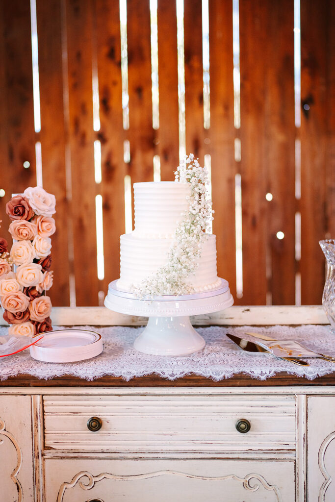 Simple white two-tier wedding cake decorated with delicate baby’s breath flowers displayed on a lace-covered table against a wooden barn wall.