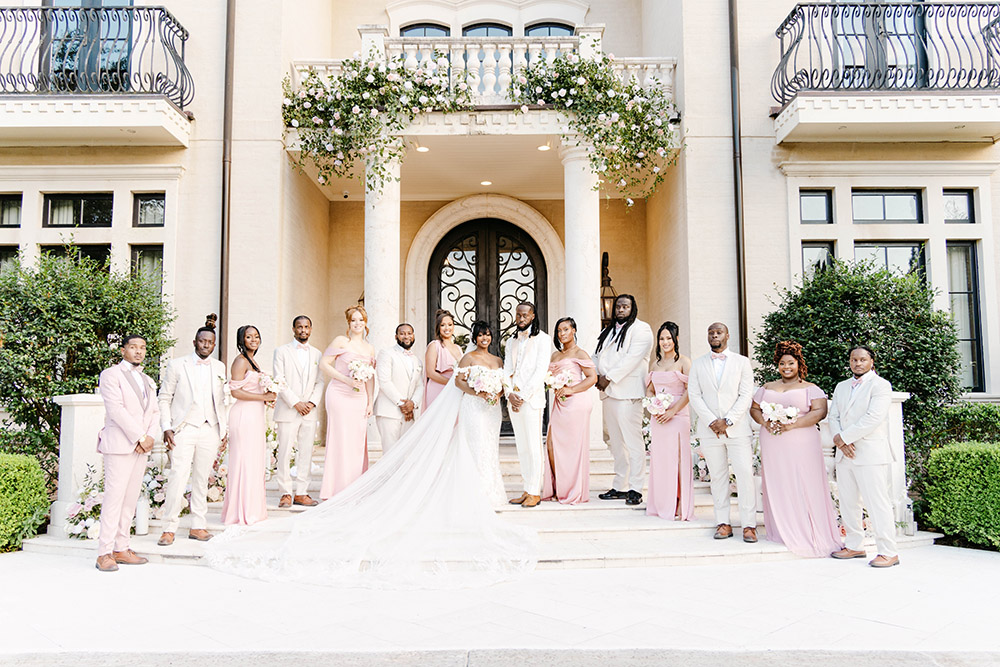 Bride and groom stand with their wedding party on marble steps in coordinated blush bridesmaid dresses and ivory tuxedos.