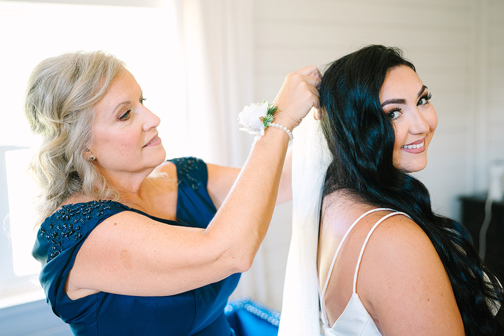 Mother of the bride adjusting the bride’s veil as the bride smiles over her shoulder while getting ready before the ceremony.