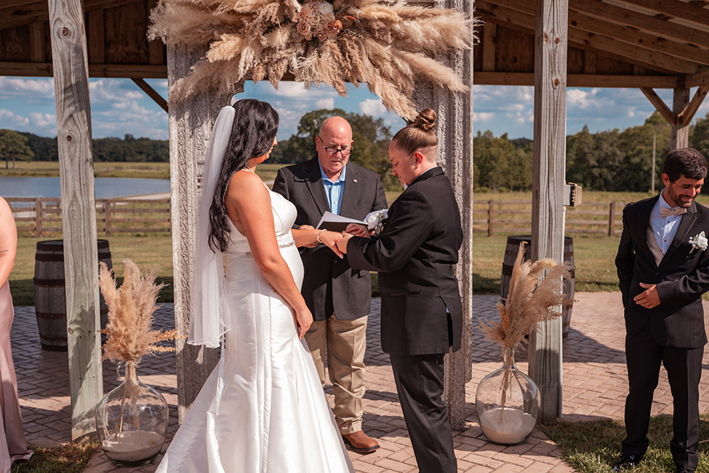 Two brides exchanging rings during an outdoor wedding ceremony beneath a pampas grass floral arch at a rustic farm venue while their officiant reads from a ceremony book.