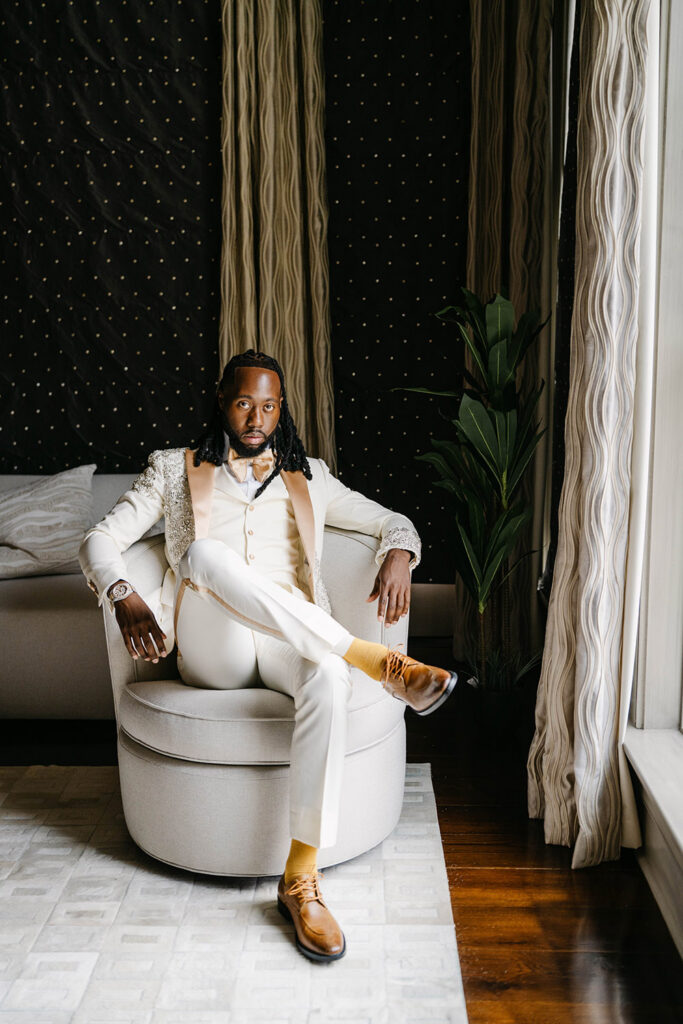 Groom in an ivory embellished tuxedo jacket sits confidently in a cream chair beside a sunlit window inside a luxury hotel suite.