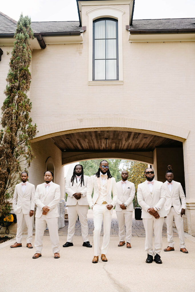 Groom and groomsmen pose beneath a grand covered entryway wearing coordinated ivory tuxedos with blush bow ties and brown dress shoes.