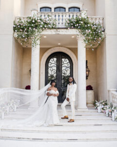 Brideโs cathedral veil billows dramatically beside the couple standing on the front steps of a floral-adorned mansion entrance.