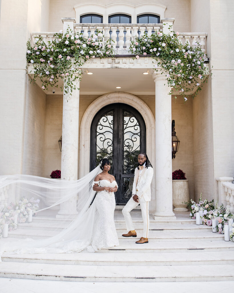 Bride’s cathedral veil billows dramatically beside the couple standing on the front steps of a floral-adorned mansion entrance.