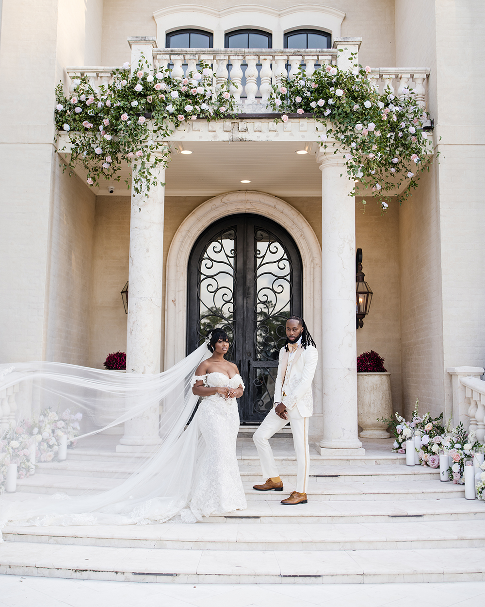 Bride’s cathedral veil billows dramatically beside the couple standing on the front steps of a floral-adorned mansion entrance.